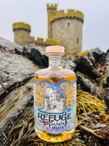 A bottle of Refuge Manx Rum sits on a rock on the coast with a castle in the background.