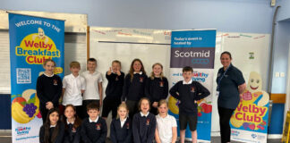 A group of kids in school sit between banners for the Healthy Living Programme Welby Breakfast Club and Scotmid Co-op.