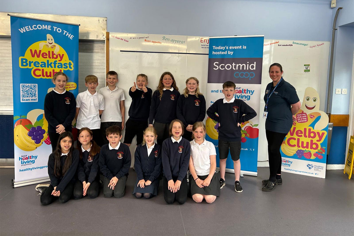 A group of kids in school sit between banners for the Healthy Living Programme Welby Breakfast Club and Scotmid Co-op with Siobhan Sandbach, Scotmid membership & community manager (North).