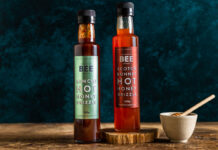 Bottles of The Scottish Bee Company Hot Honey stand on a wooden table including Kimchi Hot Honey Drizzle and Scotch Bonnet Hot Honey Drizzle with a small ceramic bowl with a honey dipper stick in it.