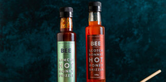 Bottles of The Scottish Bee Company Hot Honey stand on a wooden table including Kimchi Hot Honey Drizzle and Scotch Bonnet Hot Honey Drizzle with a small ceramic bowl with a honey dipper stick in it.