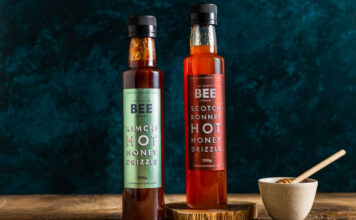 Bottles of The Scottish Bee Company Hot Honey stand on a wooden table including Kimchi Hot Honey Drizzle and Scotch Bonnet Hot Honey Drizzle with a small ceramic bowl with a honey dipper stick in it.