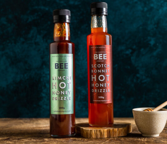 Scottish Bee Company jumps on hot honey trend Bottles of The Scottish Bee Company Hot Honey stand on a wooden table including Kimchi Hot Honey Drizzle and Scotch Bonnet Hot Honey Drizzle with a small ceramic bowl with a honey dipper stick in it.