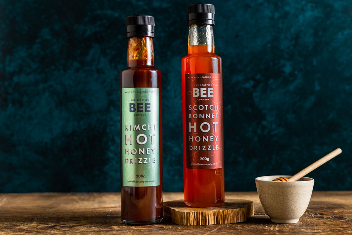 Bottles of The Scottish Bee Company Hot Honey stand on a wooden table including Kimchi Hot Honey Drizzle and Scotch Bonnet Hot Honey Drizzle with a small ceramic bowl with a honey dipper stick in it.