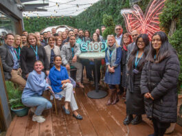 *Shopt kicks off Refer a Friend scheme A group of retailers stand together for the *Shopt Refer a Friend Scheme launch with a neon sign of the *shopt logo.