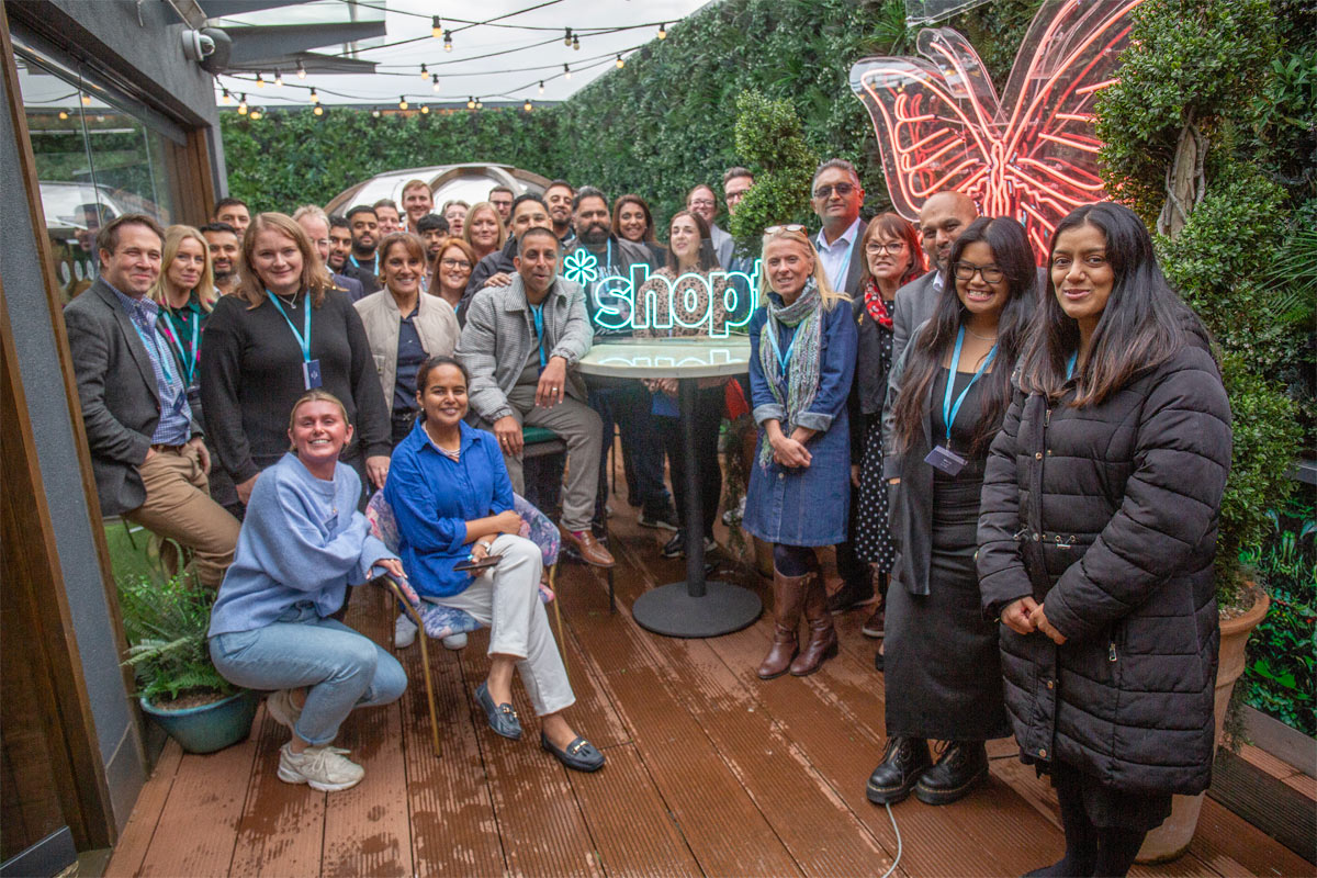 A group of retailers stand together for the *Shopt Refer a Friend Scheme launch with a neon sign of the *shopt logo.