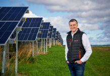 Simon Howie, owner of Simon Howie Group, stands in front of rows of solar panels.