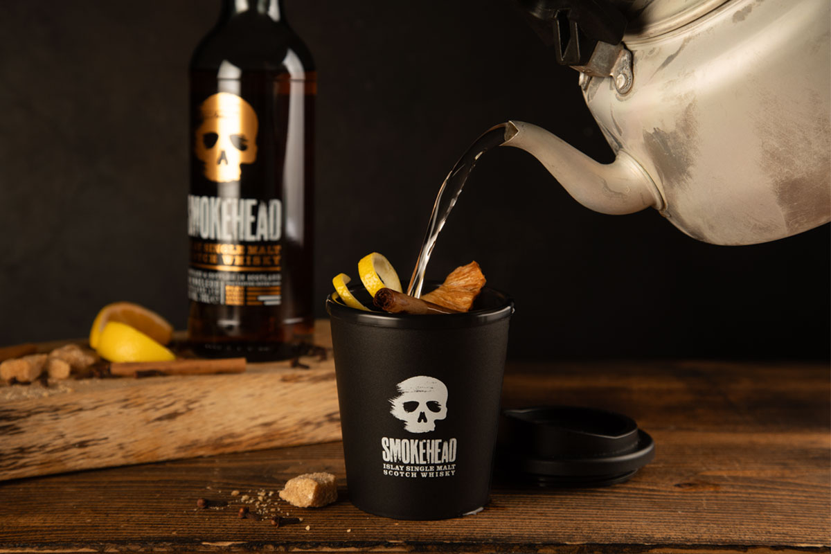 Someone pours boiling water from a kettle into a Smokehead branded cup to make a Hot Toddy cocktail with a bottle of Smokehead whisky behind it.