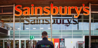 A person in a 'We hae meat' branded jumper stands in front of the entrance to a Sainsbury's supermarket.