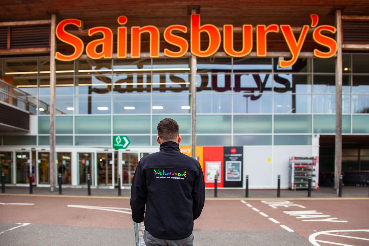 A person in a 'We hae meat' branded jumper stands in front of the entrance to a Sainsbury's supermarket.