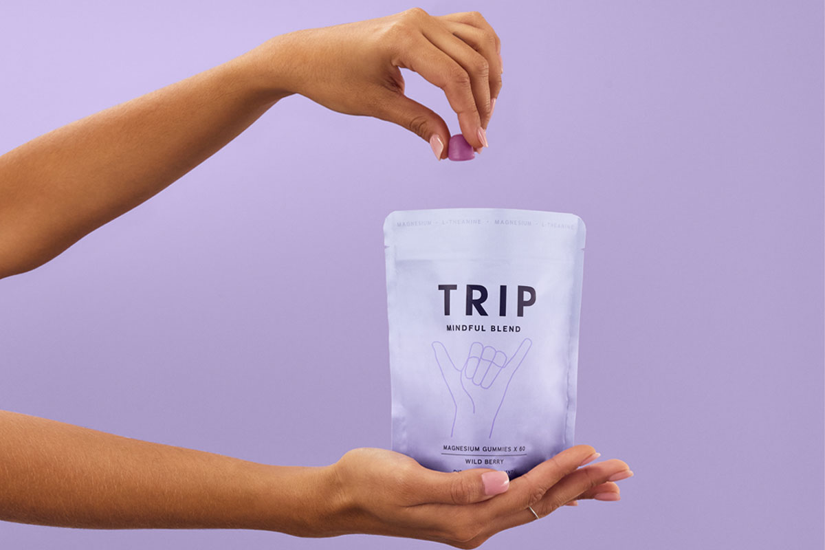 A person holds a bag of TRIP Magnesium Gummies and takes a few out with their other hand against a lilac purple background.