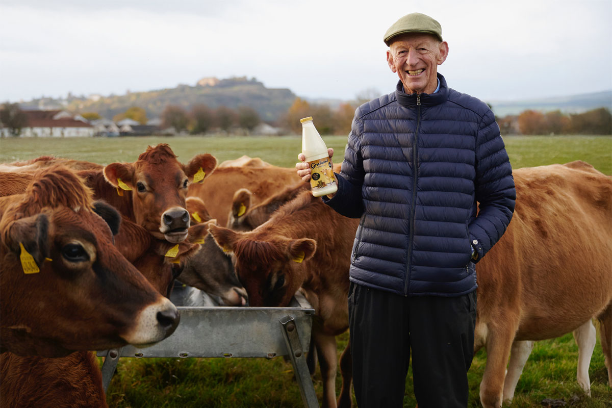 Dr Robert Graham stands in a field holding a bottle of Graham's Jersey Milk with the family's herd of cows behind him.