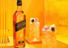 A bottle of Johnnie Walker Black Label is next to two cocktail serves set against an orange and yellow background.