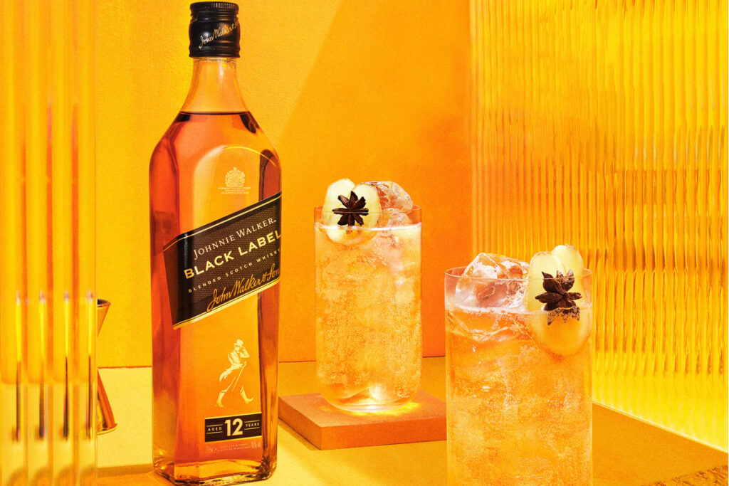 A bottle of Johnnie Walker Black Label is next to two cocktail serves set against an orange and yellow background.