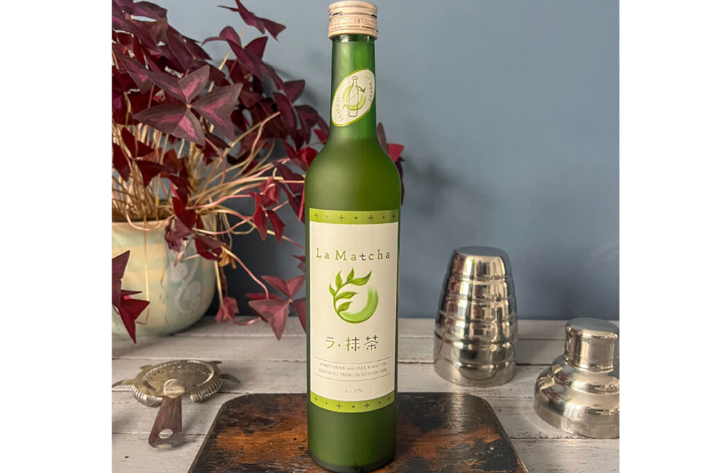 A bottle of La Liqueur Matcha sits on a table with a wooden board beneath it and a disassembled cocktail shaker to its right and a potted plant to its left.