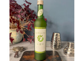 Love Drinks expands La Liqueur A bottle of La Liqueur Matcha sits on a table with a wooden board beneath it and a disassembled cocktail shaker to its right and a potted plant to its left.