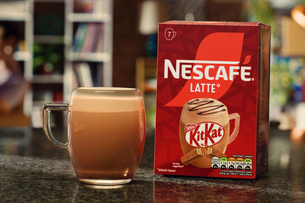 A pack of Nescafé KitKat Latte sits on a table with a cup of the coffee standing next to it.