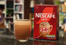 A pack of Nescafé KitKat Latte sits on a table with a cup of the coffee standing next to it.