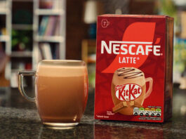 A pack of Nescafé KitKat Latte sits on a table with a cup of the coffee standing next to it.