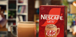 A pack of Nescafé KitKat Latte sits on a table with a cup of the coffee standing next to it.