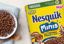 A cereal box of Nesquik Minis sits on table with a bowl filled with the cereal and a spoon next to the bowl.