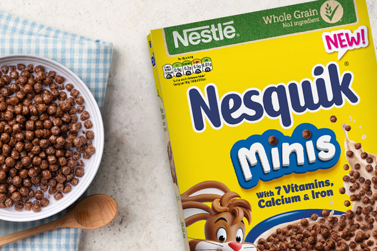 A cereal box of Nesquik Minis sits on table with a bowl filled with the cereal and a spoon next to the bowl.