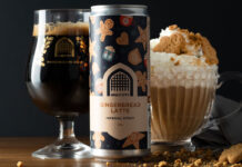 A can of Vault City Gingerbread Latte Stout stands on a table in front of a Vault City branded glass and a bowl with a gingerbread latte in it with whipped cream and gingerbread biscuit.