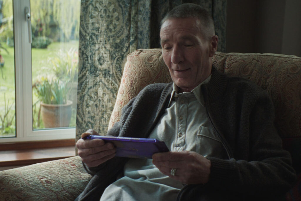 An elderly man sits in a chair by a window holding a large Cadbury Dairy Milk tablet bar.