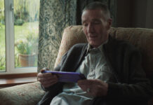 An elderly man sits in a chair by a window holding a large Cadbury Dairy Milk tablet bar.