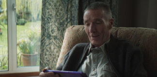 An elderly man sits in a chair by a window holding a large Cadbury Dairy Milk tablet bar.