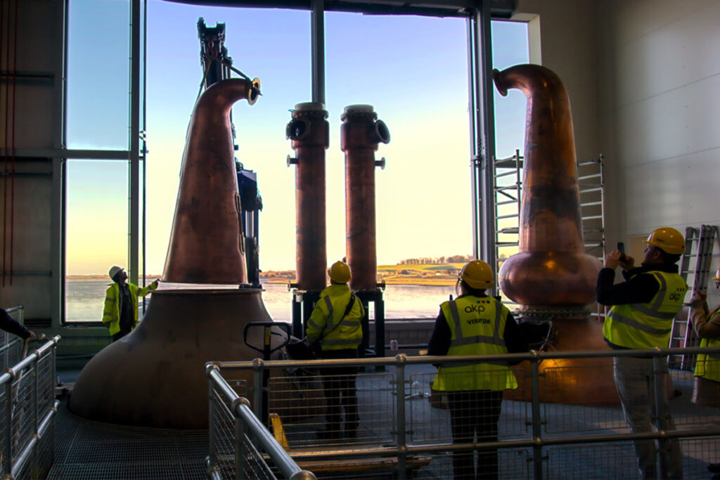 Stills being installed at Eden Mill Distillery with a large window in the background overlooking a waterfront landscape.