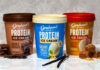 Packs of Graham's Protein Ice Creams sit on a countertop including Double Chocolate, Creamy Vanilla and Salted Caramel variants, with pieces of chocolate next to the Double Chocolate; vanilla pods next to the Creamy Vanilla; and pieces of caramel next to the Salted Caramel.