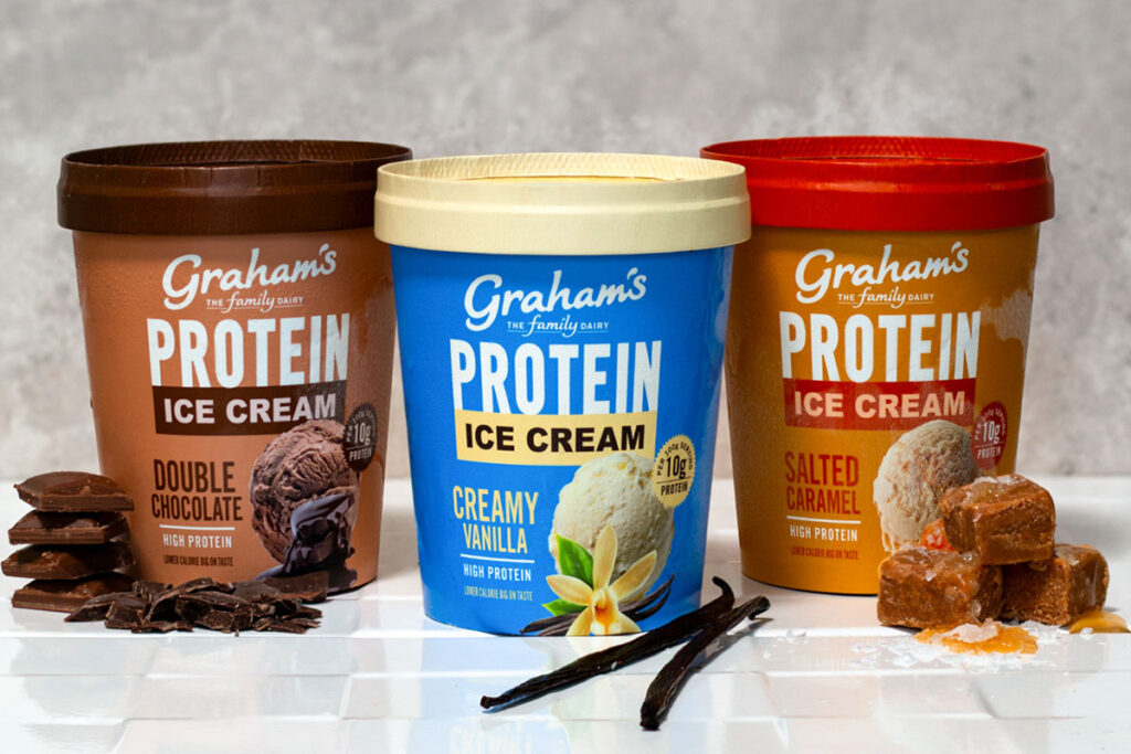 Packs of Graham's Protein Ice Creams sit on a countertop including Double Chocolate, Creamy Vanilla and Salted Caramel variants, with pieces of chocolate next to the Double Chocolate; vanilla pods next to the Creamy Vanilla; and pieces of caramel next to the Salted Caramel.