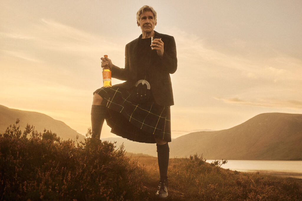 Hollywood actor Harrison Ford stands against a Highlands landscape during sunset in highland attire holding a bottle of Glenmorangie in one hand and a glass with the whisky inside it in the other.