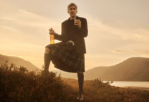 Hollywood actor Harrison Ford stands against a Highlands landscape during sunset in highland attire holding a bottle of Glenmorangie in one hand and a glass with the whisky inside it in the other.