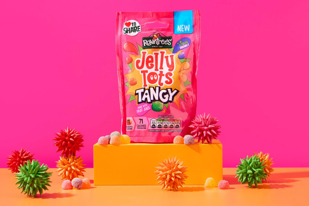 A packet of Jelly Tots Tangy stands on an orange platform against a pink background with spiky balls of different colours around the platform.