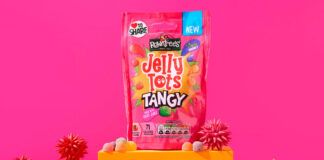 A packet of Jelly Tots Tangy stands on an orange platform against a pink background with spiky balls of different colours around the platform.