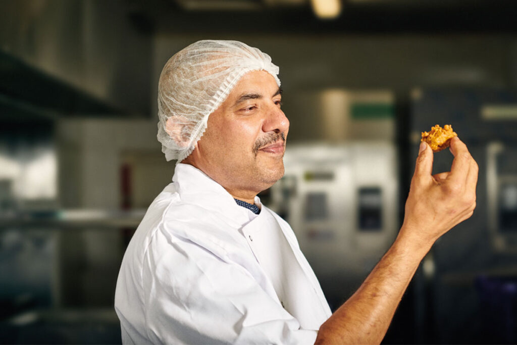 A chef holds a piece of Punjab Pakora Square Sausage Pakora in his hand looking at it.