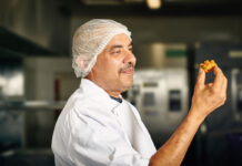 A chef holds a piece of Punjab Pakora Square Sausage Pakora in his hand looking at it.