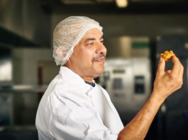 A chef holds a piece of Punjab Pakora Square Sausage Pakora in his hand looking at it.