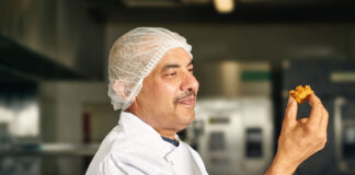 A chef holds a piece of Punjab Pakora Square Sausage Pakora in his hand looking at it.