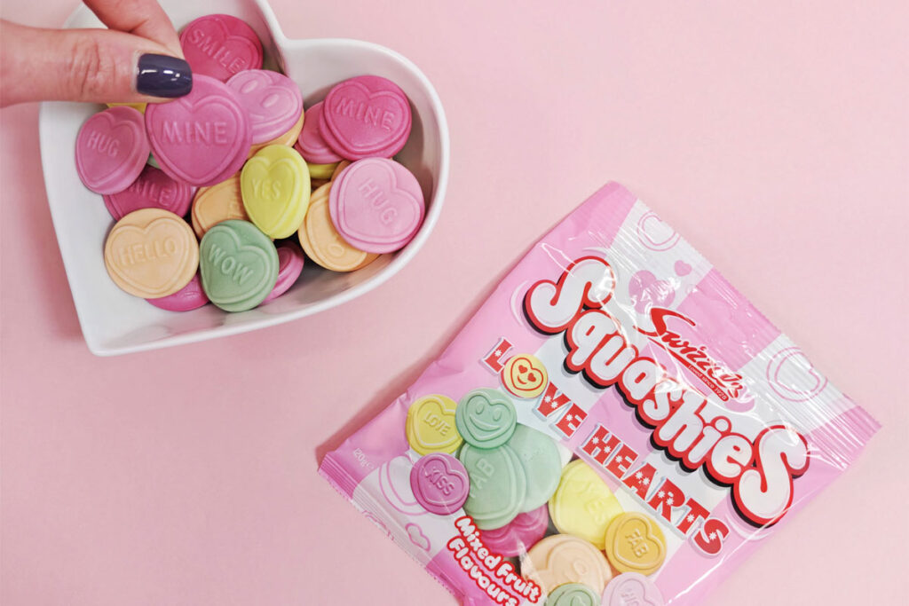 A packet of Swizzels Squashies Love Hearts lies on a pink background with a heart-shaped bowl next to it with the sweets decanted into the bowl as a hand picks one out.