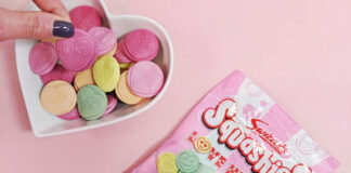 A packet of Swizzels Squashies Love Hearts lies on a pink background with a heart-shaped bowl next to it with the sweets decanted into the bowl as a hand picks one out.