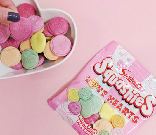 Swizzels spreads the Love with new Squashies variant A packet of Swizzels Squashies Love Hearts lies on a pink background with a heart-shaped bowl next to it with the sweets decanted into the bowl as a hand picks one out.