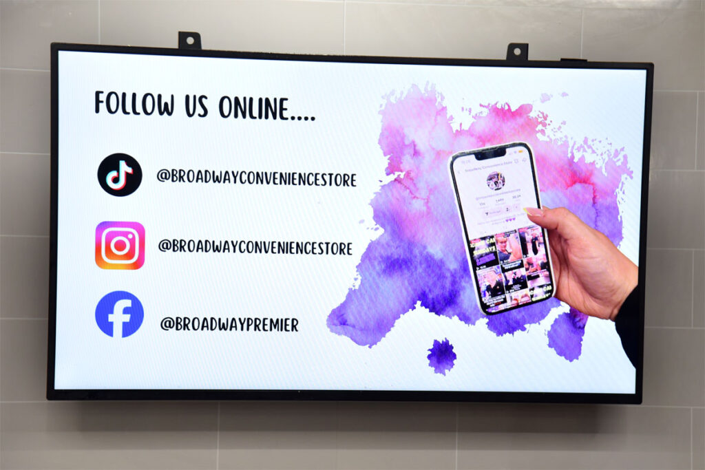 An in-store TV screen featuring social media channel links for Premier Broadway Convenience Store including TikTok, Instagram and Facebook with a hand holding a phone showing the store's TikTok page.