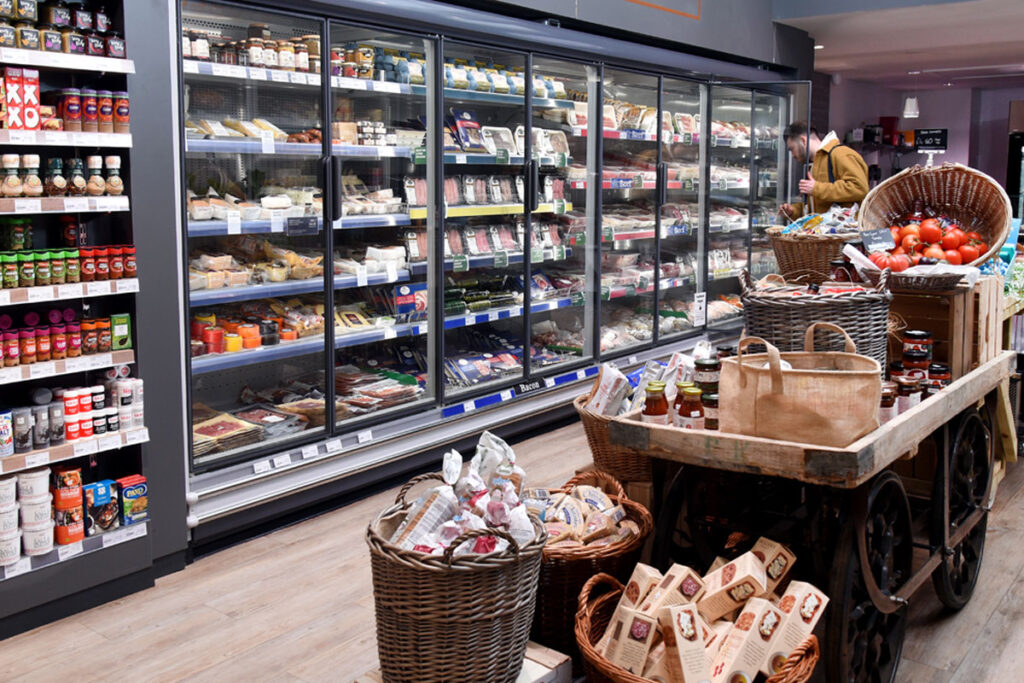 A range of chillers inside a store stocked up with meat products with a stand in the middle of the floor with some cupboard essentials on them.