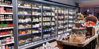 A range of chillers inside a store stocked up with meat products with a stand in the middle of the floor with some cupboard essentials on them.
