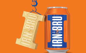 A can of Irn-Bru stands against an orange background with golden Irn-Bru Girder design hangs next to it, suspended by a drawing of a crane.