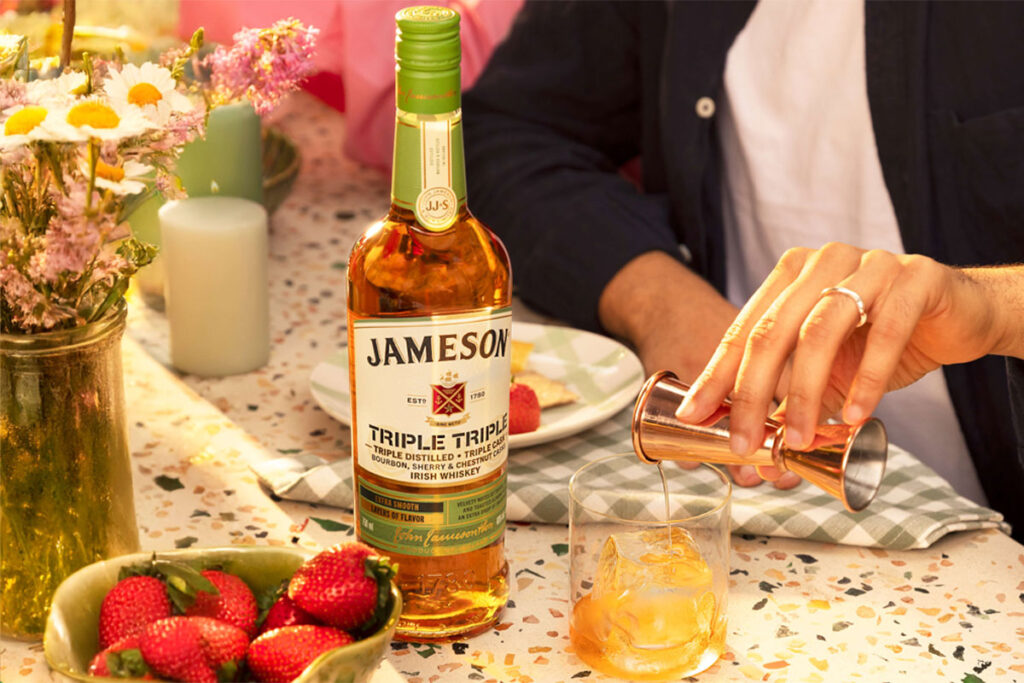 A bottle of Jameson Triple Triple Chestnut Edition is on a table as a person pours out a measure of the whiskey from a jigger into a glass with ice with a vase filled with flowers on the table and a bowl of strawberries.