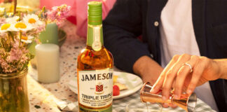 A bottle of Jameson Triple Triple Chestnut Edition is on a table as a person pours out a measure of the whiskey from a jigger into a glass with ice with a vase filled with flowers on the table and a bowl of strawberries.